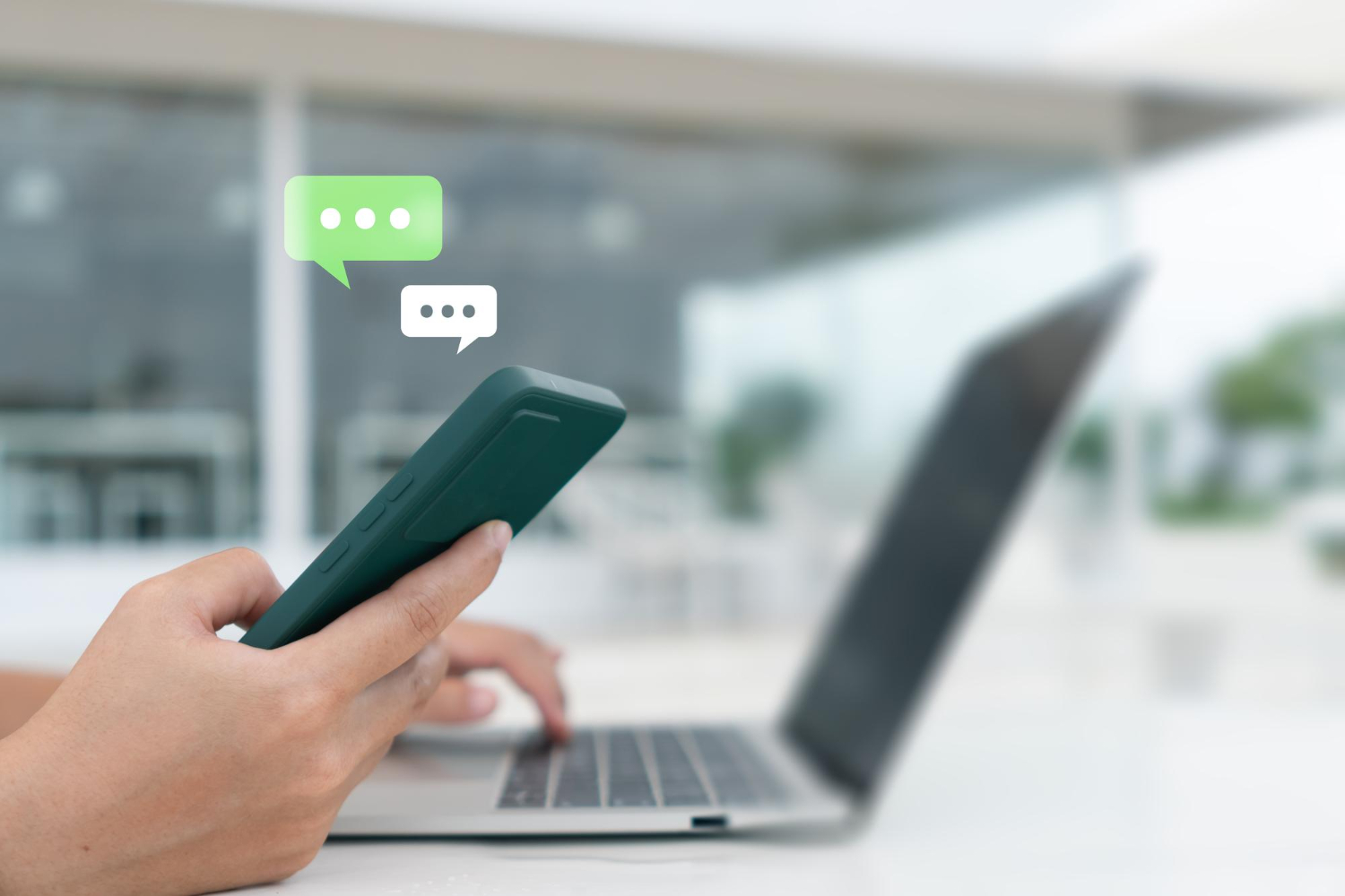 A person holds a smartphone with a laptop in front of them; two chat message icons appear above the phone, illustrating the ease of SMS Marketing communication.