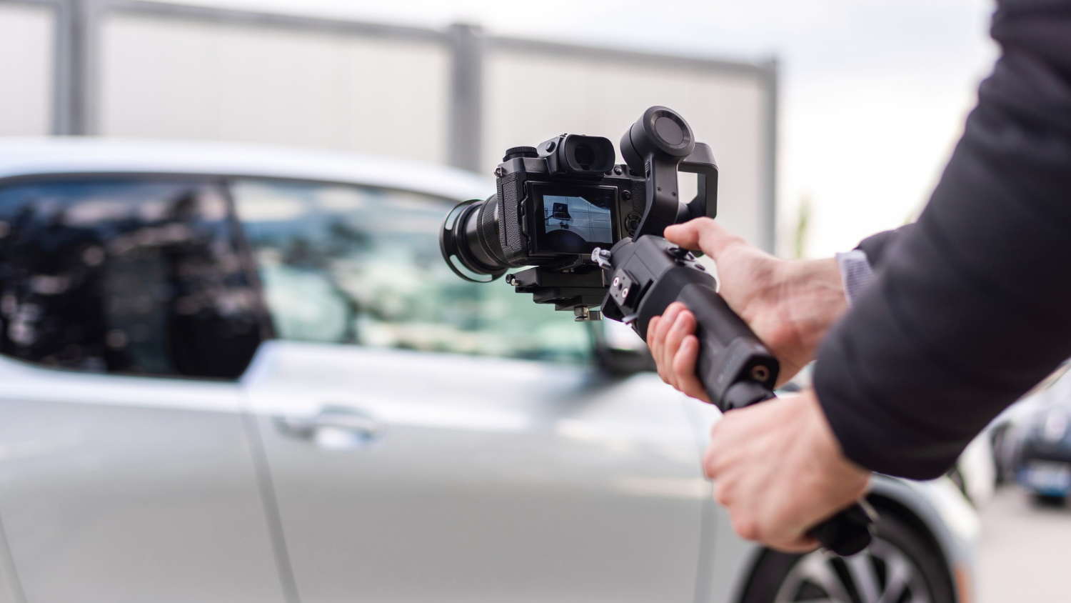 A person holding a camera mounted on a stabilizer is filming a silver car parked outside, capturing the moment for video or photography projects.