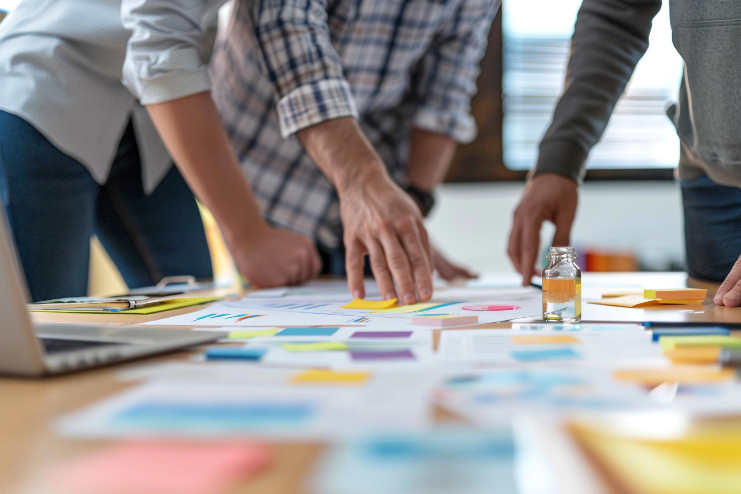 Three people stand around a table, reviewing documents and charts as they collaborate on a marketing strategy, with various papers, sticky notes, and a small bottle visible in the foreground.