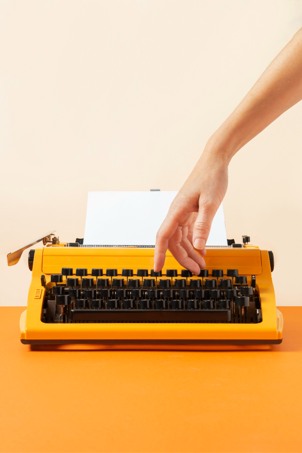 A hand reaches to type on a yellow typewriter with a blank sheet of paper, ready for content writing, on an orange surface and a beige background.