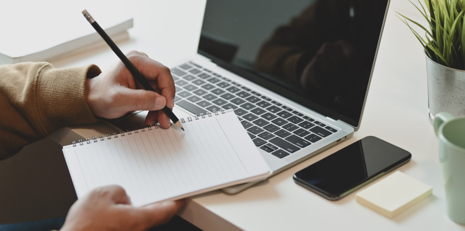 Person engaged in content writing, jotting notes in a notebook beside a laptop, smartphone, sticky notes, and a green cup on a white desk.