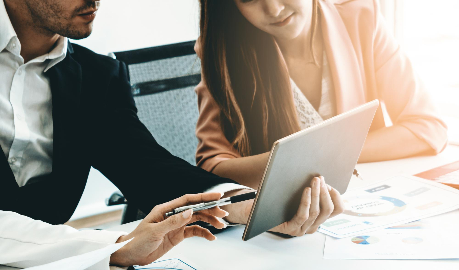 Two people in business attire review data on a tablet at a desk with printed charts and papers.