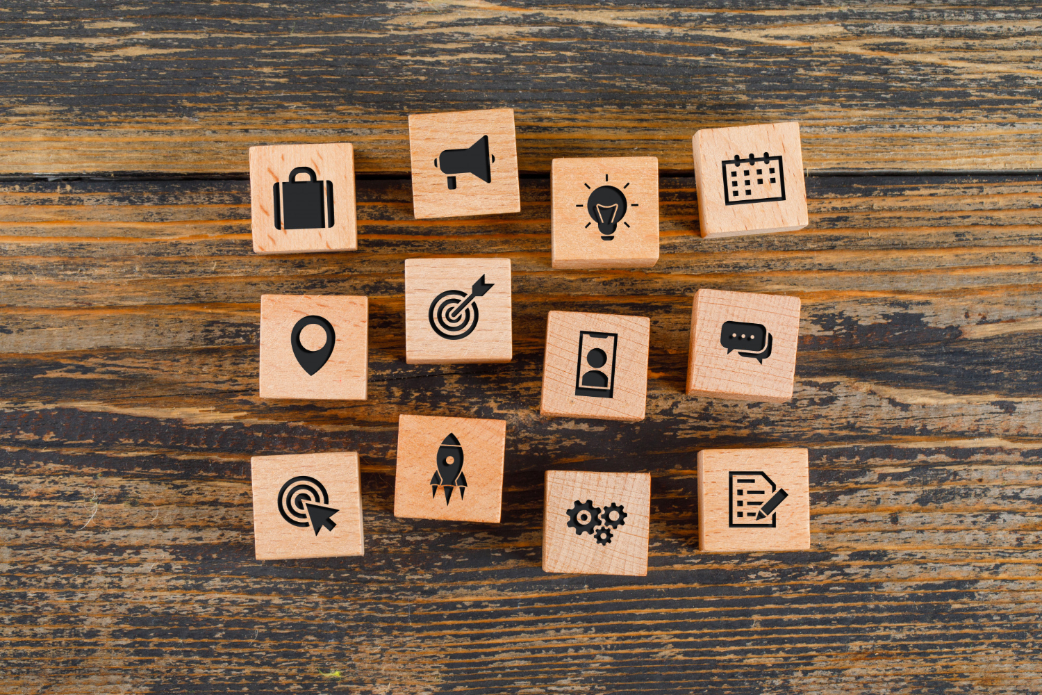 Wooden blocks with black icons representing business and productivity concepts—like a briefcase, target, strategy gears, rocket, checklist, calendar, and speech bubble—arranged on a wooden surface to illustrate marketing strategy ideas.