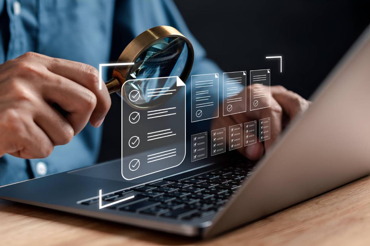 A person holds a magnifying glass over a laptop keyboard with digital checklists and charts displayed, symbolising data analysis, compliance check or online review - reflecting how SMS marketing could be the next big thing in the future of marketing.