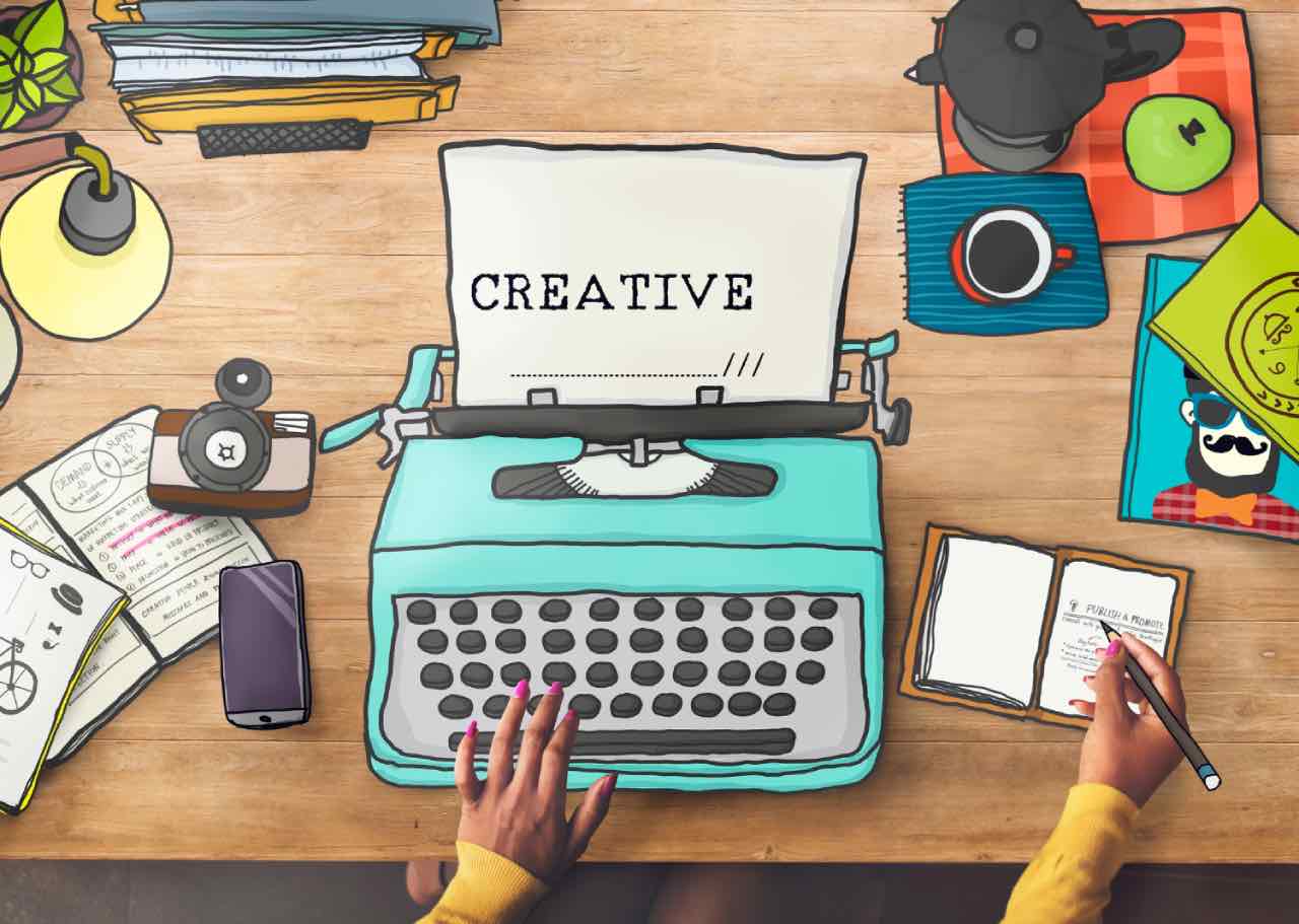 A person types "CREATIVE" on a blue typewriter at a cluttered wooden desk filled with papers, notebooks, a camera, coffee, and a phone - crafting social media content that connects ideas and inspires.