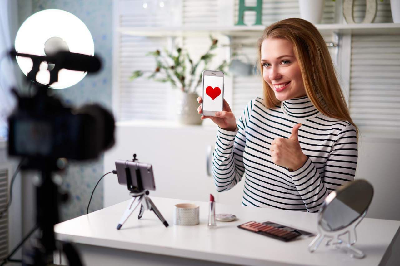 A woman sits at a desk with makeup items, giving a thumbs up and holding a phone displaying a heart, showcasing how social proof affects buyers decision making on online shopping.