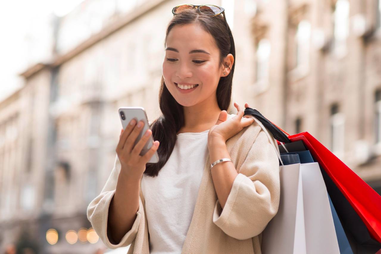 A woman is smiling while looking at her smartphone and holding several shopping bags on a city street, excited about the latest SMS marketing offers and imagining the future of shopping.