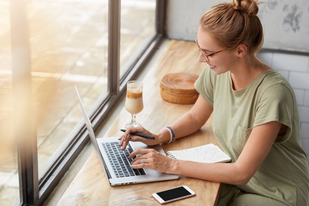 Woman wearing glasses uses a laptop at a wooden table, holding a pen and notebook perfectly poised to elevate skills in content writing, with a smartphone and iced coffee nearby.