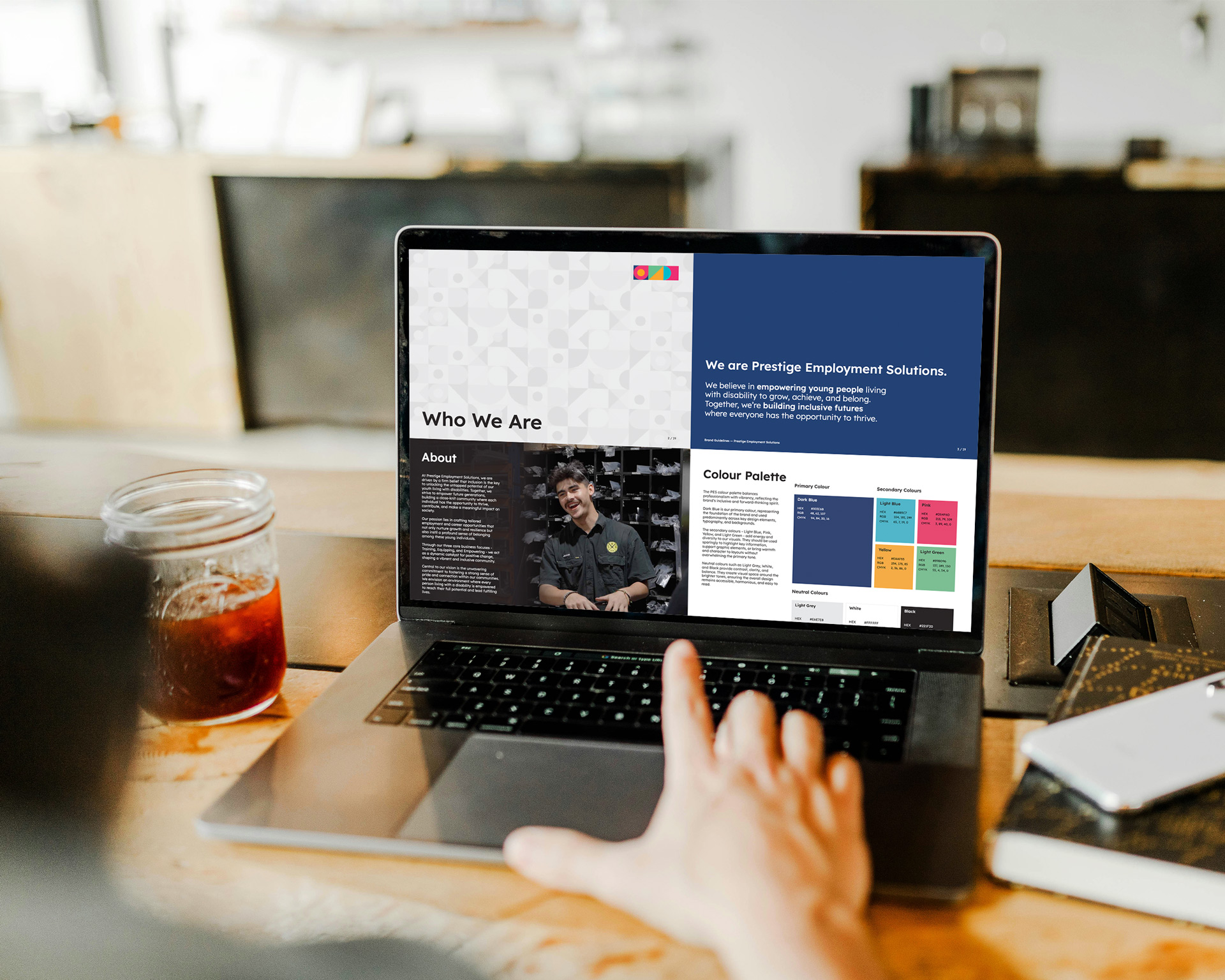 A person points at a laptop screen displaying a company profile webpage with sections like "Who We Are," "About," a photo of a man, and a 2026 color palette reflecting the brand identity. A drink sits next to the laptop.