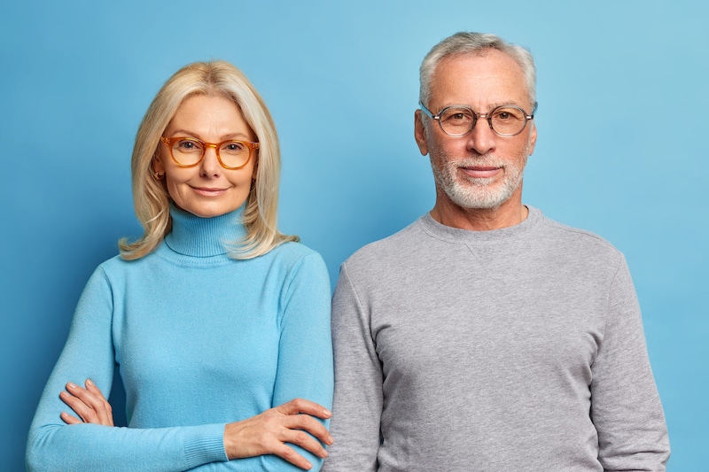         Senior couple in glasses standing against a blue background at home.